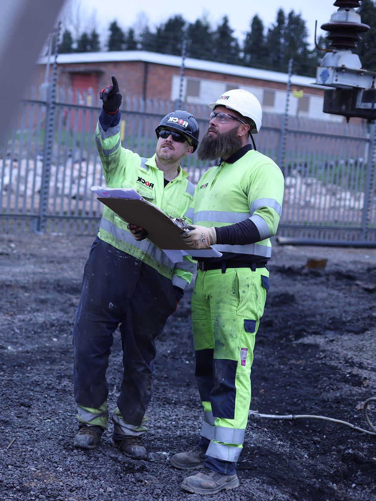 Two male 岩石电源连接 employees in high vis clothing, looking at plans and pointing upwards